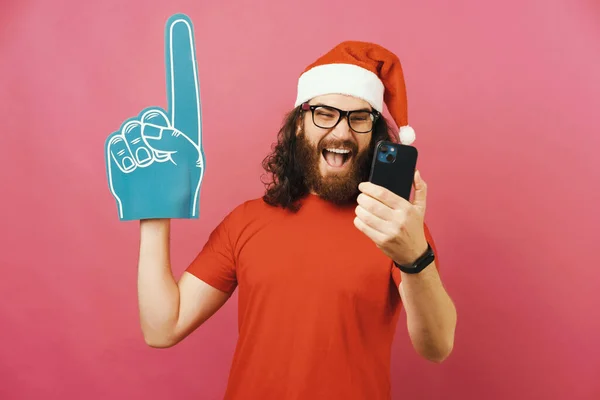 陽気な男は ファングローブを身に着けている間 携帯電話上のクリスマスサッカーの試合を見ている スタジオショットオーバーピンクの背景 — ストック写真