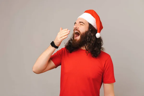 Fiatal Szakállas Férfi Ásít Miközben Egy Karácsonyi Sapkát Visel Szürke — Stock Fotó