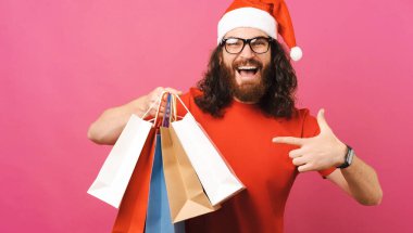 Gözlüklü ve Noel şapkalı kıvırcık adam pembe bir stüdyoda elinde tuttuğu bazı alışveriş çantalarını işaret ediyor..
