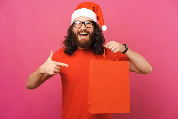 クリスマスの帽子をかぶった興奮した男が彼が持っている買い物袋を指している スタジオショットオーバーピンクの背景 — ストック写真