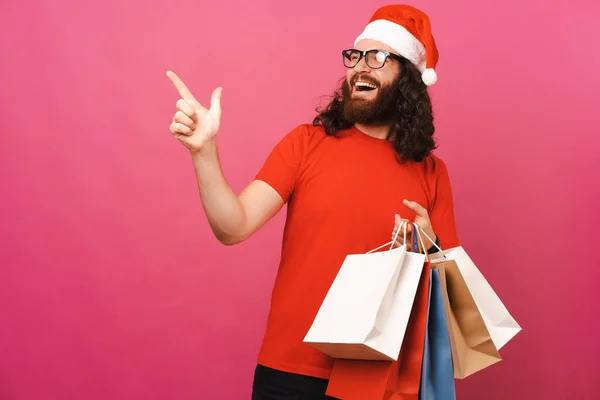 Cool Man Wearing Christmas Hat Pointing Aside Holding Shopping Bags — Stock Photo, Image