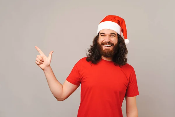 턱수염을 곱슬곱슬 남자가 크리스마스 모자를 카메라 앞에서 웃으면서 가리키고 — 스톡 사진
