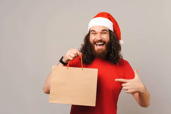 クリスマスの帽子をかぶった興奮した髭の男がクラフトショッピングバッグを指している彼は保持している — ストック写真
