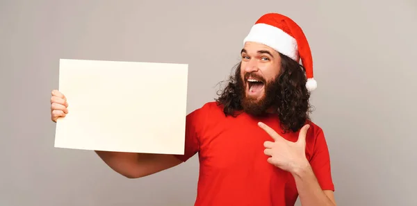 ひげを生やした男は灰色の背景の上にクリスマスに白い長方形の紙の形で保持し ポイント — ストック写真