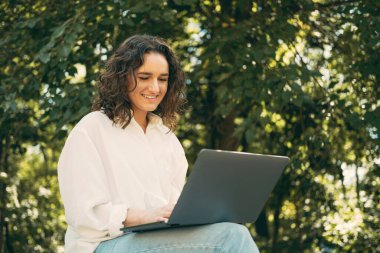 Gülümseyen kıvırcık saçlı genç bir kadının fotoğrafı bankta oturuyor ve dışarıda dizüstü bilgisayarla çalışıyor..