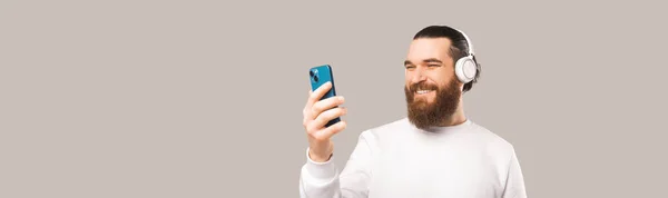 一个留着胡子的男人拿着耳机看着电话的横幅照片 在浅灰背景下的工作室拍摄 — 图库照片