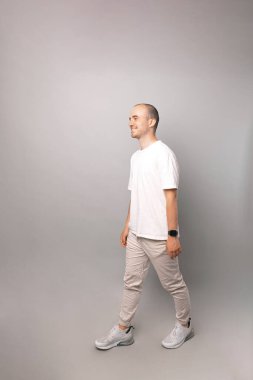 Beyaz giyen kel bir adamın dikey görüntüsü ve gri arka planda bir stüdyoda yürümesi..