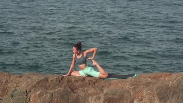 Mujer joven hace yoga en postura de paloma sobre roca grande contra mar — Vídeo de stock