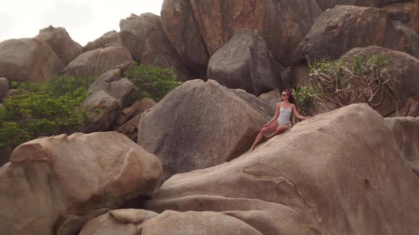 Młoda kobieta pozuje siedząc na wielkim kamieniu na dzikiej plaży — Wideo stockowe