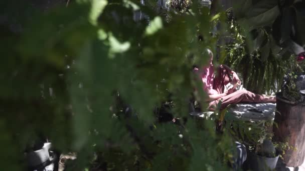 Mann liest Buch auf Bank hinter dicken Bäumen im Garten — Stockvideo