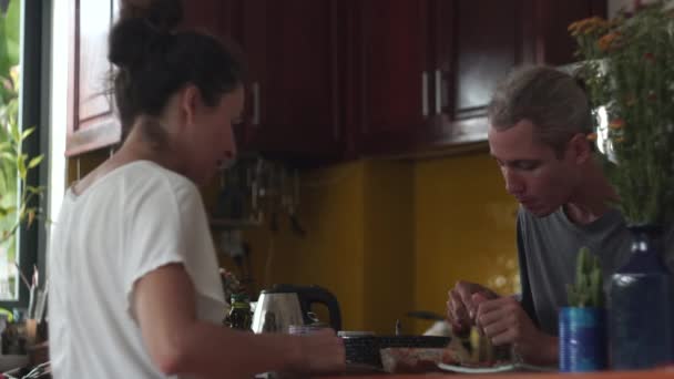 Junger Mann und Frau essen und trinken sitzend in kleiner Küche — Stockvideo