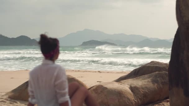 Giovane donna si siede sulla sabbia della spiaggia selvaggia guardando il mare infuria — Video Stock