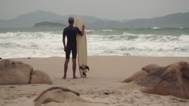Hombre con tabla de surf mira las olas del mar de pie en la playa — Vídeos de Stock