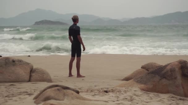Серфер дивиться на бурхливі хвилі, що стоять на піщаному пляжі — стокове відео