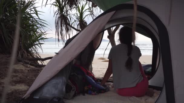 Девушка сидит в палатке на диком пляже и смотрит на море — стоковое видео