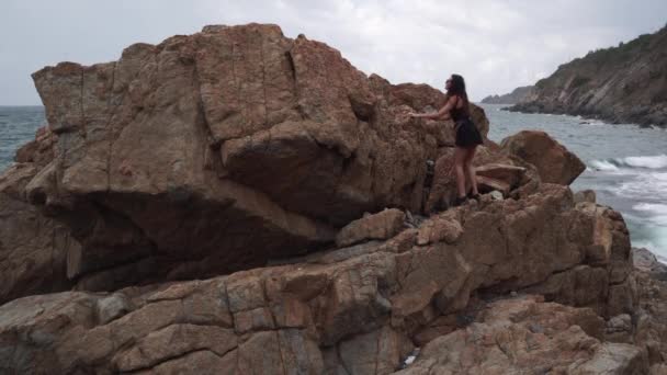 Turista em vestido preto sobe em grande penhasco velho na praia — Vídeo de Stock