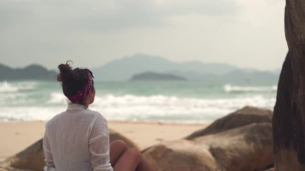 Молода жінка сидить біля скель, дивлячись на море на дикому пляжі — стокове відео
