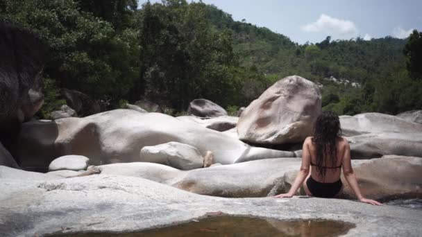 Frau sitzt auf riesigem Fluss-Felsen gegen Waldhügel — Stockvideo