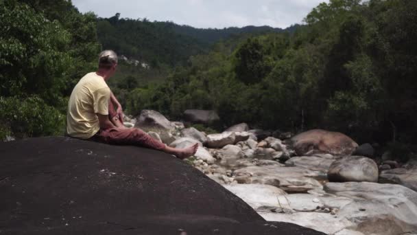 El hombre mira montañas forestales sentadas en roca por río — Vídeos de Stock