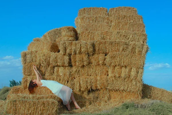 a young woman is sitting on a haystack in a field with a straw hat.