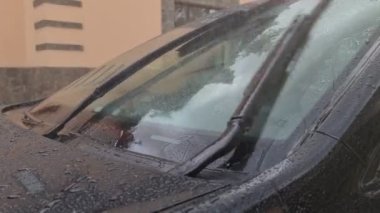 Siyah bir arabanın ön camına yağmur yağar. Silecekler çalışıyor. Cam silecekleri yağmuru temizler..