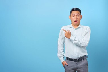 Şok olmuş genç yakışıklı işadamı mavi arka plandaki fotokopi alanını işaret ediyor