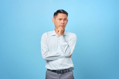 Düşünceli genç ve yakışıklı işadamı çenesini ovuşturuyor ve mavi arka planda bir soru düşünüyor