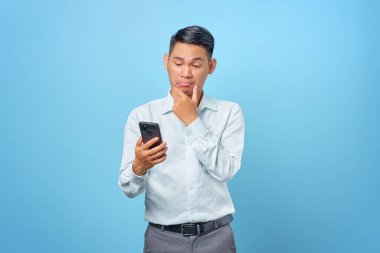 Dalgın genç iş adamı mavi arka planda akıllı telefon kullanıyor.