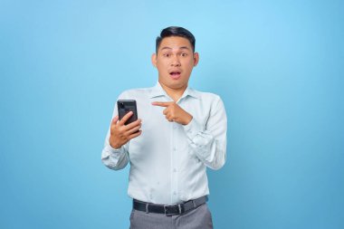 Şok olmuş genç yakışıklı işadamı mavi arka plandaki akıllı telefonu işaret ediyor.