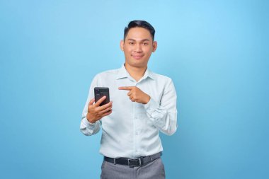 Gülümseyen yakışıklı iş adamı mavi arka plandaki akıllı telefonu işaret ediyor.