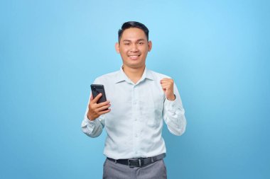 Akıllı telefon tutan neşeli, genç ve yakışıklı iş adamı mavi arka planda kazanan jestler yapıyor.