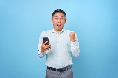 Heyecanlı genç yakışıklı iş adamı elinde akıllı telefon ile mavi arka planda kazanan jestler yapıyor.