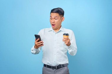 Şok olmuş genç yakışıklı iş adamı elinde akıllı telefon ve mavi arka planda bir fincan kahve tutuyor.