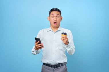 Şok olmuş genç yakışıklı iş adamı elinde akıllı telefon ve mavi arka planda bir fincan kahve tutuyor.