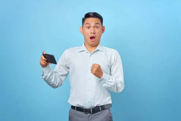 Şok olmuş genç ve yakışıklı iş adamı elinde akıllı telefonla mavi arka plandaki kameraya bakıyor.