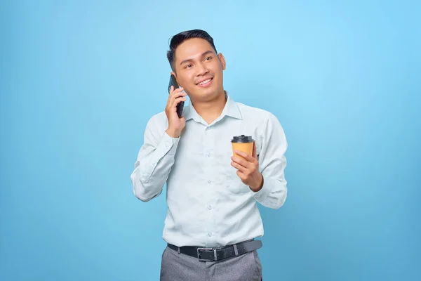 Gülümseyen yakışıklı iş adamı akıllı telefondan konuşuyor ve mavi arka planda bir fincan kahve tutuyor.