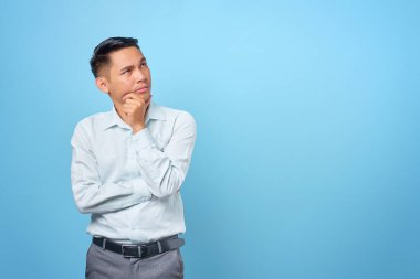 Düşünceli genç ve yakışıklı işadamı çenesini ovuşturuyor ve mavi arka planda bir soru düşünüyor