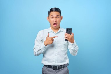 Şok olmuş genç iş adamının portresi mavi arka plandaki akıllı telefonu işaret ediyor.