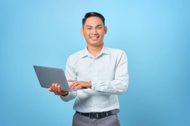 Gülümseyen yakışıklı iş adamı elinde dizüstü bilgisayarla mavi arka plandaki kameraya bakıyor.