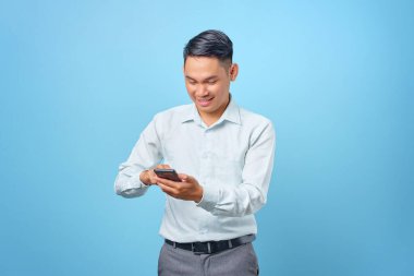 Gülümseyen yakışıklı iş adamı mavi arka planda akıllı telefonu kullanıyor ve mesaj atıyor.