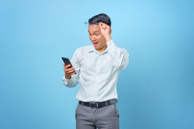 Şok olmuş genç ve yakışıklı iş adamı akıllı telefon kullanıyor ve mavi arka planda gözlüklerini çıkarıyor.