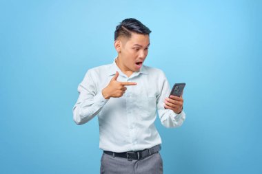 Şok olmuş genç yakışıklı işadamı mavi arka plandaki akıllı telefonu işaret ediyor.