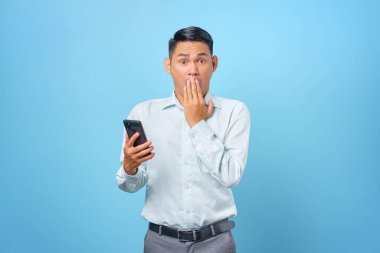 Şok olmuş genç yakışıklı iş adamı elinde akıllı telefon ve mavi arka planda ağzını kapatıyor.