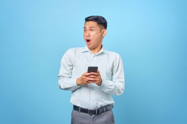 Şok olmuş genç ve yakışıklı iş adamı elinde akıllı bir telefonla mavi arka plana bakıyor.