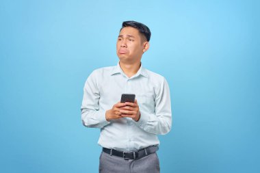 Üzgün, genç, yakışıklı iş adamı elinde akıllı bir telefonla mavi arka plana bakıyor.