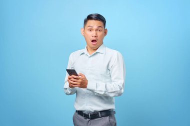 Şok olmuş genç ve yakışıklı iş adamı elinde akıllı telefonla mavi arka plandaki kameraya bakıyor.