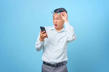 Şok olmuş genç ve yakışıklı iş adamı akıllı telefon kullanıyor ve mavi arka planda gözlüklerini çıkarıyor.