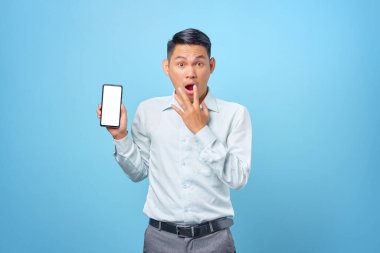 Şok olmuş genç yakışıklı iş adamı mavi arka planda akıllı telefon boş ekranı gösteriyor.
