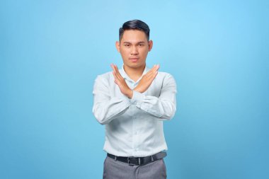 Ciddi, genç, yakışıklı iş adamı elini çapraz yapıp mavi arka planda reddetme hareketi yapıyor.