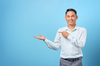 Gülümseyen genç ve yakışıklı iş adamı mavi arka planda avuçlarında kopya alanı gösteriyor